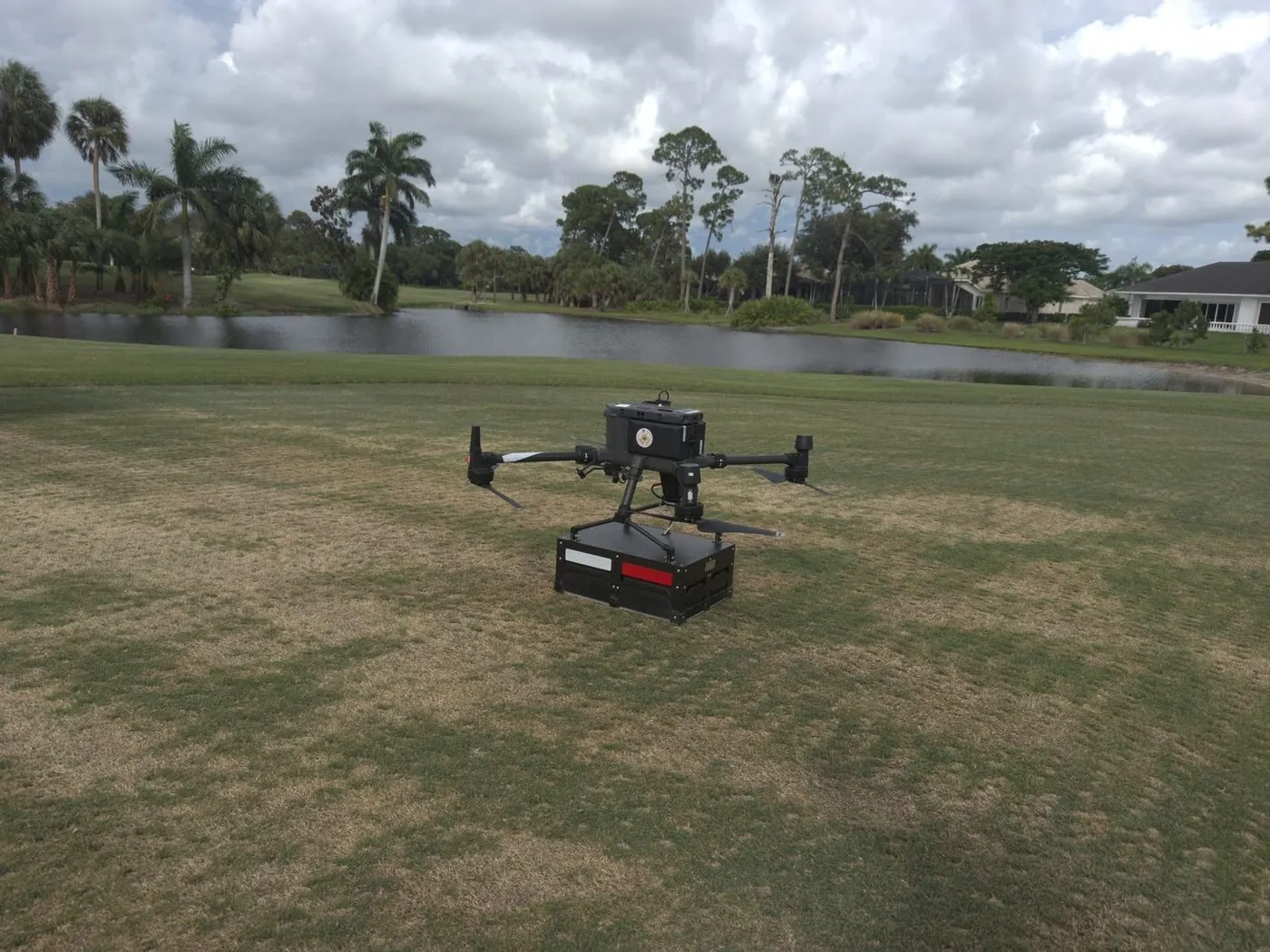 DJI M300 RTK with Zond Aero GPR antenna hovering low over golf course turf beside a water hazard, surveying for buried irrigation lines